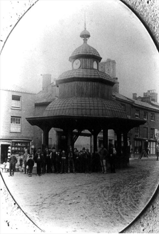 Photograph. North Walsham Market Cross, c1880's. Dyball, Earthenware Dealer on left....sold shop to Stead & Simpson's around 1890. (North Walsham Archive).